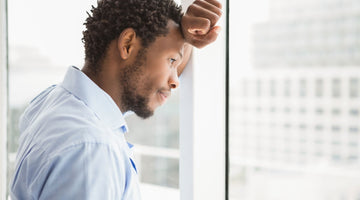 Man wearing blue collard shirt leaning on window looking out into the distance from a highrise office building